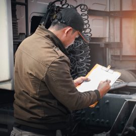 operador de camión revisando documentación enfrente del larguero de un tractocamión, con chamarra café, seguro de equipo pesado #Qualitas